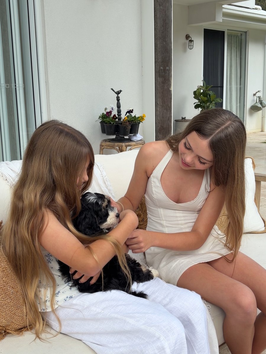 Two girls on an outdoor couch nursing a Bernedoodle puppy between them